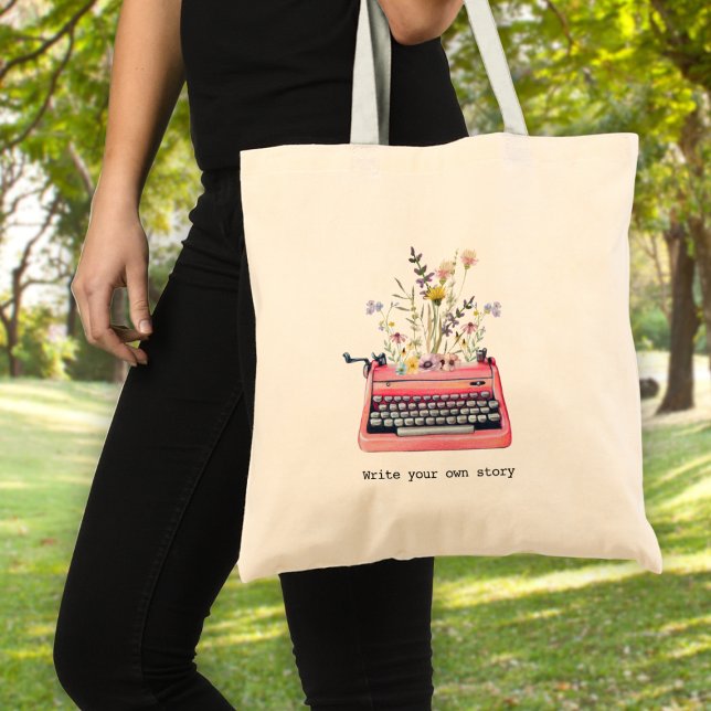 Bolsa Tote Escreva sua própria história, Escritores (A woman holding a tote bag with a typewriter graphic and wildflowers & text: "write your own story")