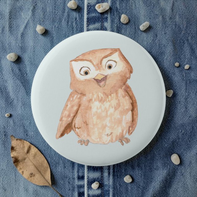 Bóton Redondo 5.08cm Coruja Feliz Aquarela (A watercolor owl cartoon smiling on a pale blue pin, laid on denim amid pebbles and leaves.)