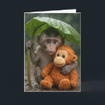 Cartão Thinking of You Macaque Monkey with Orangutan<br><div class="desc">A young macaque monkey uses a large green leaf as a makeshift umbrella while holding onto a stuffed orange orangutan toy in the gentle rain of a lush forest.
Inside text can be changed.</div>