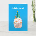 Cupcake do cartão de Natal (Galês) com árvore de<br><div class="desc">Um cartão de Natal brilhante e alegre de Galês com uma árvore de Natal decorada sobre um cupcake congelado, de uma colagem de papel pintado à mão por Judy Adamson. O cumprimento dianteiro, no texto branco em um fundo azul, é "Nadolig Llawen!" e a mensagem interna, que você pode mudar,...</div>