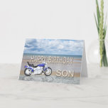 Filho, um cartão de aniversário com uma bicicleta<br><div class="desc">Um ciclo de motor azul que está em uma praia com as nuvens macias no céu. O feliz aniversario escrito na rocha refletiu na água que encontra-se na areia.</div>