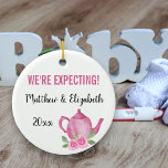 Ornamento De Cerâmica Esperamos que uma menina esteja criando bule<br><div class="desc">Bonito ornamento para sua gravidez que você pode personalizar. Um dos lados tem flores cor-de-rosa e tipografia escrita escrita com letra de mão que diz "Uma menina está se preparando". O outro lado tem um bule rosa fofo e seu texto personalizado. O modelo está configurado para que você edite o...</div>