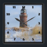 Relógio Quadrado Farol resistente numa costa rochosa no inverno<br><div class="desc">Um farol robusto de tijolos vermelhos fica sozinho protegendo os mares no inverno. O chão áspero e rochoso está coberto de neve com gramíneas coloridas de trigo soprando suavemente no vento. Evoca uma sensação de solidão. Um céu azul com nuvens brancas e espertas.</div>