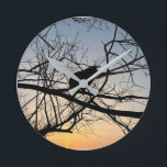 Relógio Redondo Golden Sunset Silhouette: Bird on Tree Branches<br><div class="desc">Capture the serene beauty of twilight with this stunning silhouette photograph. A lone bird rests among the intricate web of bare tree branches, set against a vibrant golden and blue sunset sky. The high contrast and natural geometry create a peaceful, artistic atmosphere that complements any modern decor. This design is...</div>